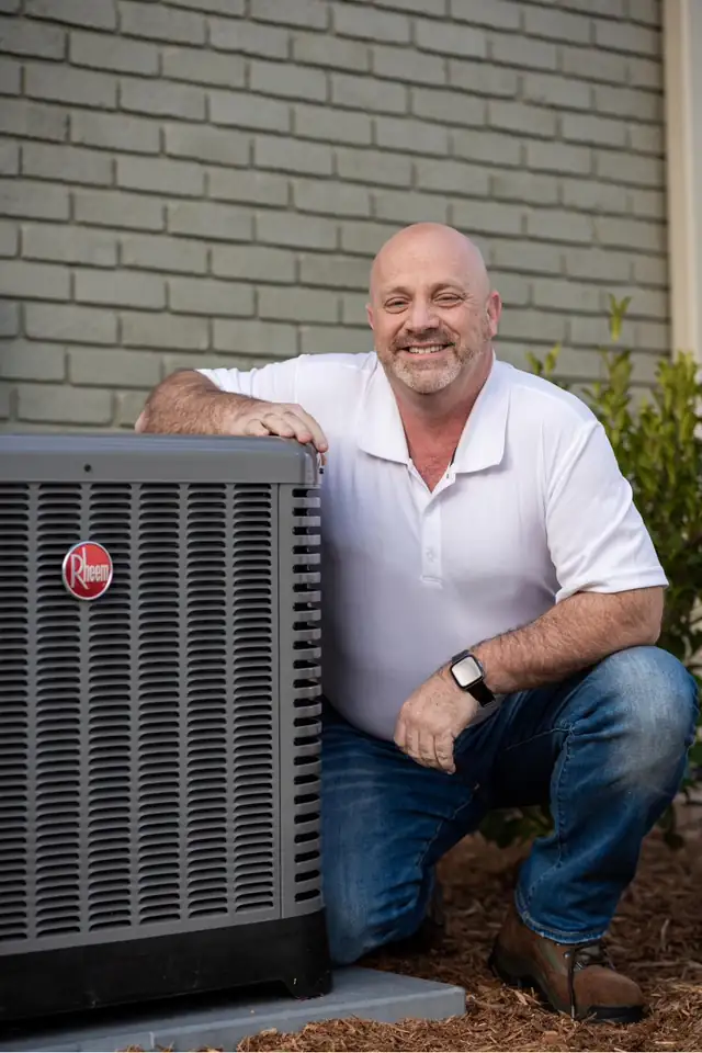 Shane & Son's technician kneeling beside a Rheem outdoor air conditioning unit